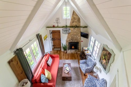 Cozy living room with a high ceiling, stone fireplace, red sofa, patterned chairs, and natural light from large windows.