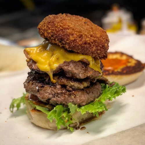 A towering burger with three beef patties, melted cheese, lettuce, and a crispy top bun, served on a toasted bottom bun.