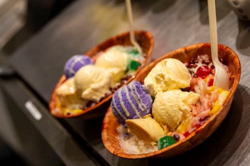 Two bowls of colorful ice cream topped with fruits and jellies, served with spoons on a dark surface.