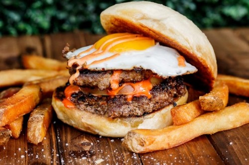 A juicy burger with a fried egg, spicy sauce, and two patties, served with crispy fries on a wooden table.
