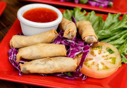 A plate of crispy spring rolls on a bed of purple cabbage, with a side of dipping sauce and a slice of tomato.