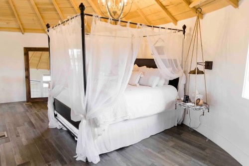 A cozy bedroom featuring a four-poster bed draped in sheer white curtains, with wooden ceilings and stylish decor.