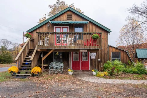 A rustic wooden house with a green roof, red doors, and a porch adorned with flowers and seating.
