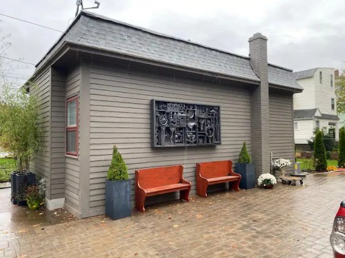 A gray house with a decorative wall art piece, two wooden benches, and potted plants on a rainy day.