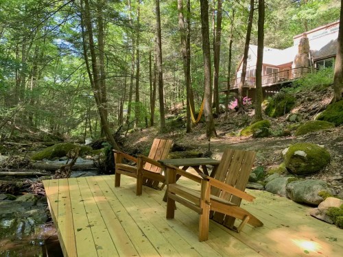 Two wooden chairs on a deck by a stream, surrounded by lush green trees and a cozy cabin in the background.