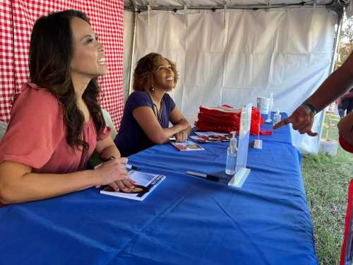 Two women smile at a table with promotional materials, while a person gestures towards them in a tented area.