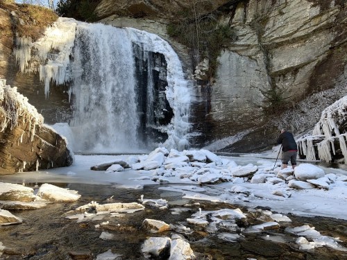 A person stands on icy rocks near a frozen waterfall, surrounded by snow and icicles in a winter landscape.
