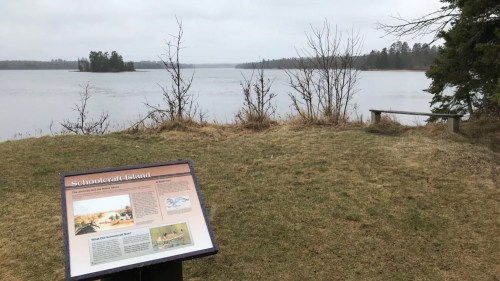 A view of a calm lake with a sign about Schoolcraft Island in the foreground, surrounded by bare trees and grass.