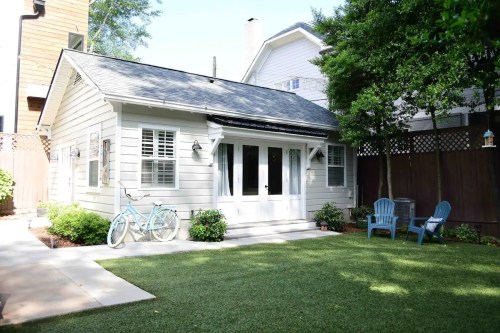 A cozy backyard with a small white cottage, green lawn, and blue chairs, surrounded by trees and plants.