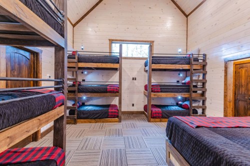 A cozy bunk room with wooden bunk beds, plaid bedding, and warm wooden walls, featuring ample natural light.