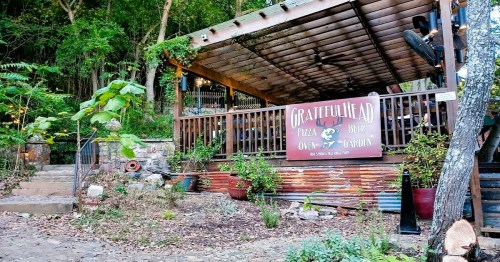 A rustic outdoor pizza restaurant with a wooden deck, surrounded by greenery and colorful plants.