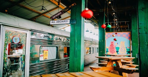A colorful diner interior featuring a vintage train, festive decorations, and wooden picnic tables.