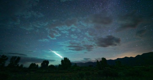 A starry night sky with clouds and a bright meteor streaking across, illuminating the landscape below.