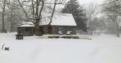 A cozy cabin surrounded by snow-covered trees, with a snowy pathway leading to a porch and chairs.