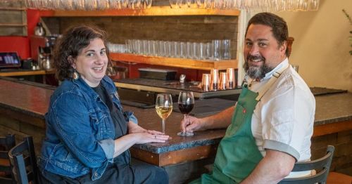 A woman and a man sit at a bar, smiling and holding glasses of wine, with a cozy atmosphere in the background.