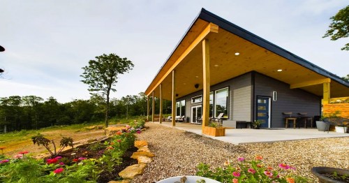 A modern house with a sloped roof, wooden pillars, and a landscaped garden featuring colorful flowers.