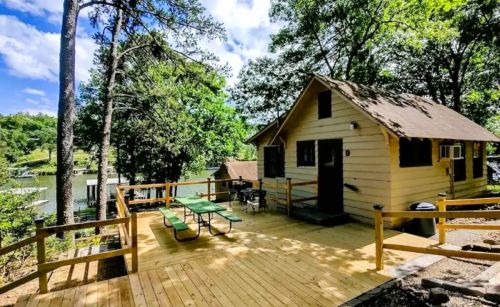 A cozy cabin by the lake with a wooden deck, picnic table, and surrounded by trees under a blue sky.