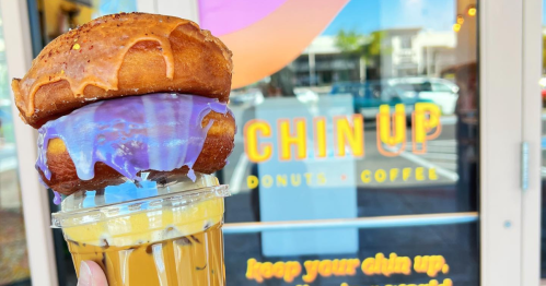 A hand holding a colorful donut sandwich above a coffee cup, with a cheerful shop window in the background.