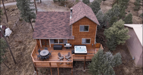 Aerial view of a wooden house with a deck, hot tub, and outdoor seating, surrounded by trees.