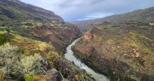 A winding river flows through a rugged canyon surrounded by green and rocky hills under a cloudy sky.