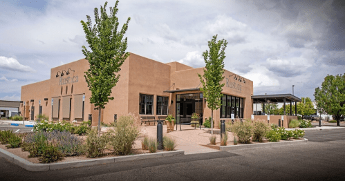 A modern adobe-style building with large windows, surrounded by greenery and a parking lot under a cloudy sky.