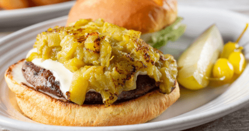 A close-up of a burger topped with green chili, served with a pickle and yellow peppers on a white plate.