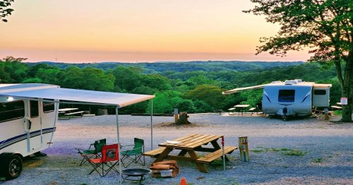A serene campsite at sunset, featuring RVs, picnic tables, and a view of lush green hills.