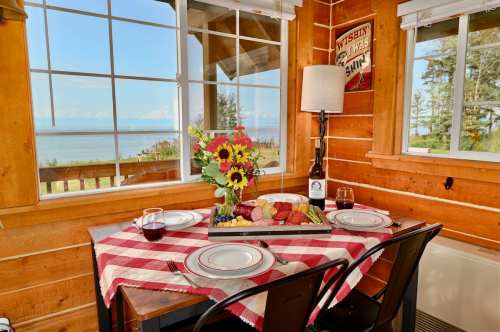 A cozy dining table with a checkered tablecloth, flowers, wine, and a scenic view of the ocean through large windows.