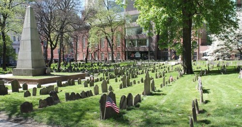 A historic cemetery with gravestones and American flags, surrounded by trees and buildings in the background.