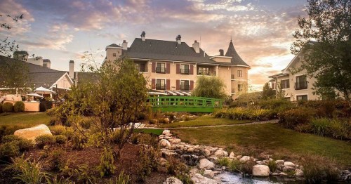 A picturesque hotel with a green bridge, surrounded by lush gardens and a serene stream at sunset.
