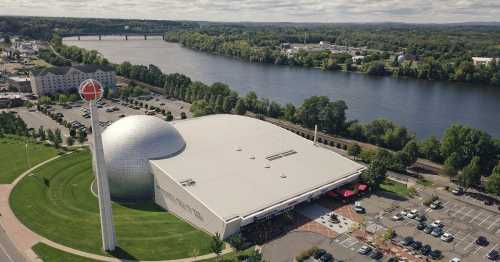 Aerial view of a modern building with a large dome, near a river and surrounded by green trees and parking spaces.