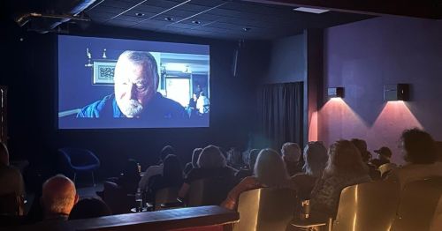 A dimly lit theater with an audience watching a film featuring a close-up of an older man's face on the screen.