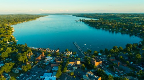 Aerial view of a serene lake surrounded by greenery and a small town, with boats and a dock visible on the water.