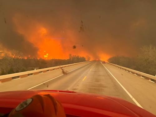 View from a vehicle on a highway, with smoke and flames visible in the distance, indicating a wildfire.