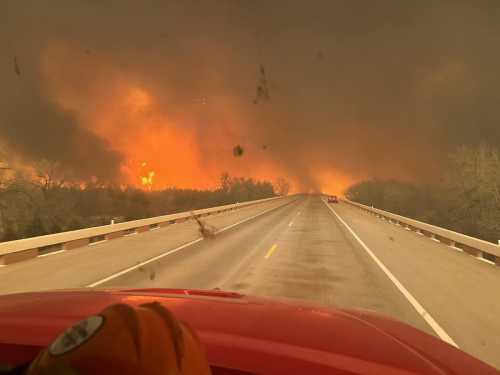 View from a vehicle on a highway, with smoke and flames visible in the distance, indicating a wildfire.