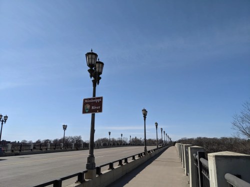 A wide view of a bridge with street lamps and a sign indicating the Mississippi River under a clear blue sky.