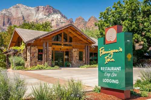 A rustic lodge with a stone exterior and wooden accents, surrounded by greenery and mountains in the background.