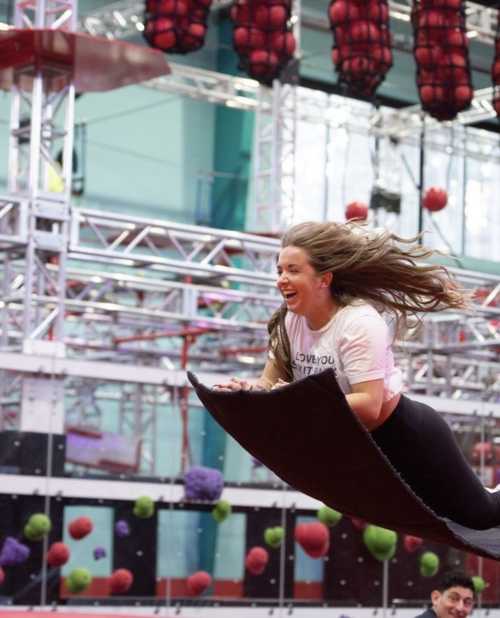 A woman joyfully flies through the air on a flying carpet-like mat in a vibrant indoor obstacle course.