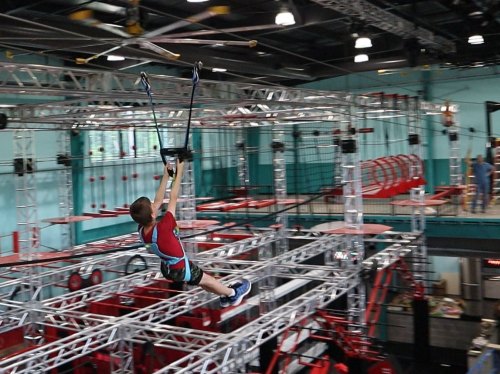 A child zip-lining across an indoor obstacle course with metal structures and various climbing elements.