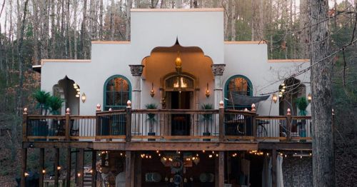 A charming, two-story house with a wooden deck, surrounded by trees and adorned with decorative lights.