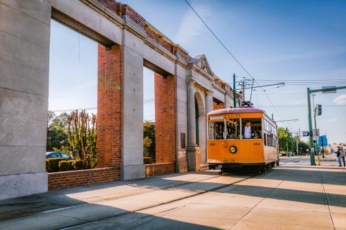 A vintage tram approaches a brick archway on a sunny day, with people walking nearby.