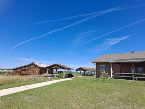 A grassy area with two wooden structures and a modern building under a clear blue sky with wispy clouds.
