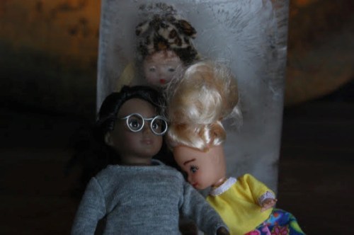 Three dolls, one with glasses, huddle together in front of a large block of ice, creating a whimsical scene.