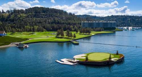 A scenic view of a golf course by a lake, surrounded by trees and mountains, with a modern building in the background.