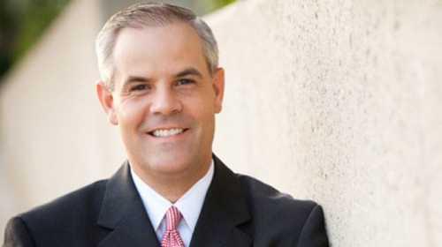 A smiling man in a suit stands against a textured wall, exuding a professional and approachable demeanor.