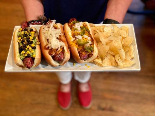 A person holds a platter with four gourmet hot dogs topped with various ingredients and a side of potato chips.