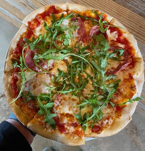 A hand holding a pizza topped with cheese, salami, and fresh arugula on a wooden table.