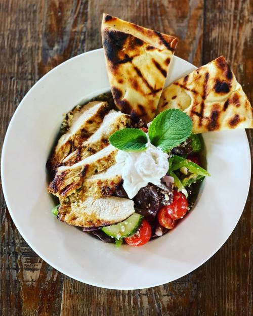 Grilled chicken salad with cherry tomatoes, olives, feta, and pita bread, garnished with mint and yogurt.