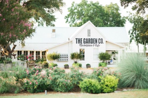 A charming white building with "The Garden Co." sign, surrounded by greenery and flowers, serving as a marketplace and café.