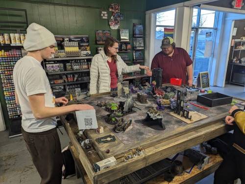A group of three people play a tabletop game at a store, surrounded by miniatures and game supplies on a large table.